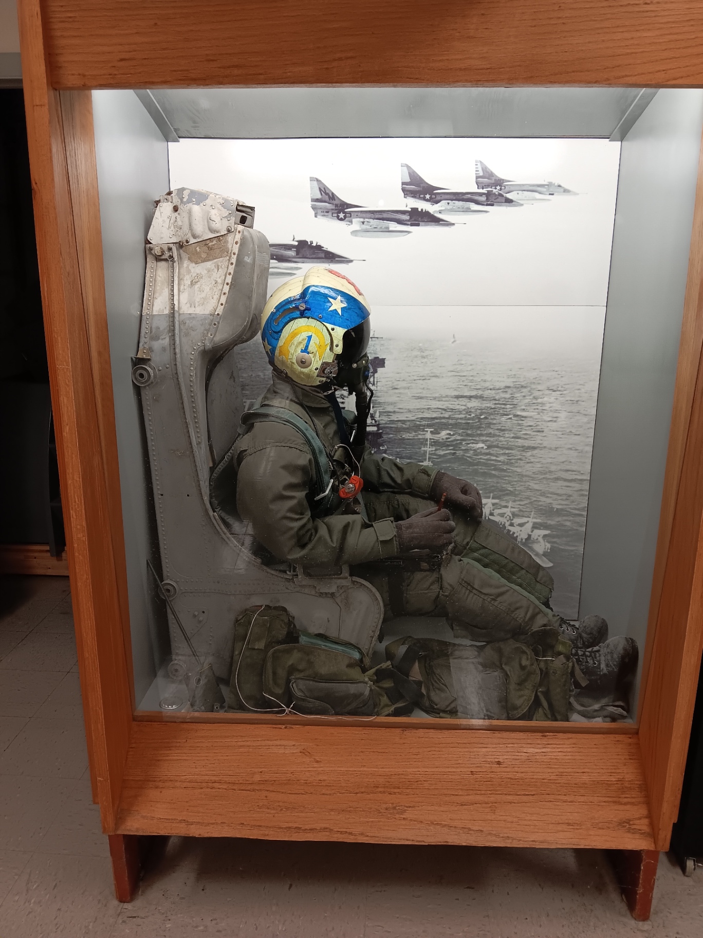 A full ejection seat display: a cadet in flight gear, helmet and oxygen mask, set against a carrier deck at sea.