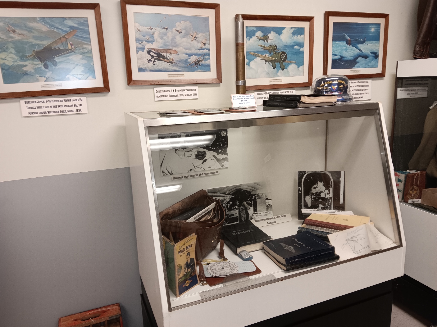 Pursuit-plane prints flown by Flying Cadet Ed Tindall in the 1930s, over a navigator cadet's case: E-6B computer, air navigation manuals and the first shell fired from the USS John Hood.