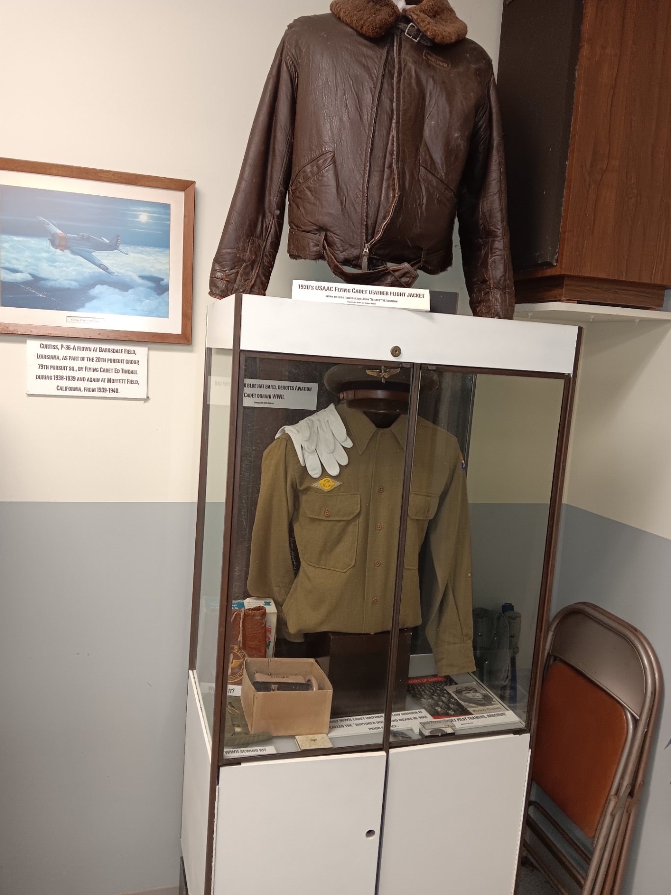 A 1930s USAAC Flying Cadet leather flight jacket above a WWII cadet uniform case — wool shirt, white gloves, dark blue hat band and a wartime sewing kit.