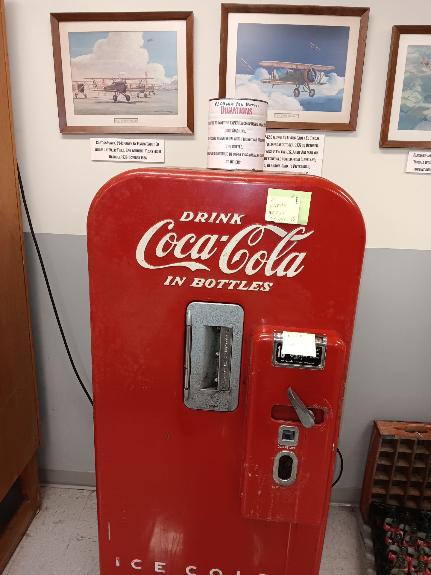 The museum's working 1940s Coca-Cola bottle machine, beneath prints of the Curtiss Hawk P1-C and Boeing P-12E flown by Flying Cadet Ed Tindall at Kelly Field.