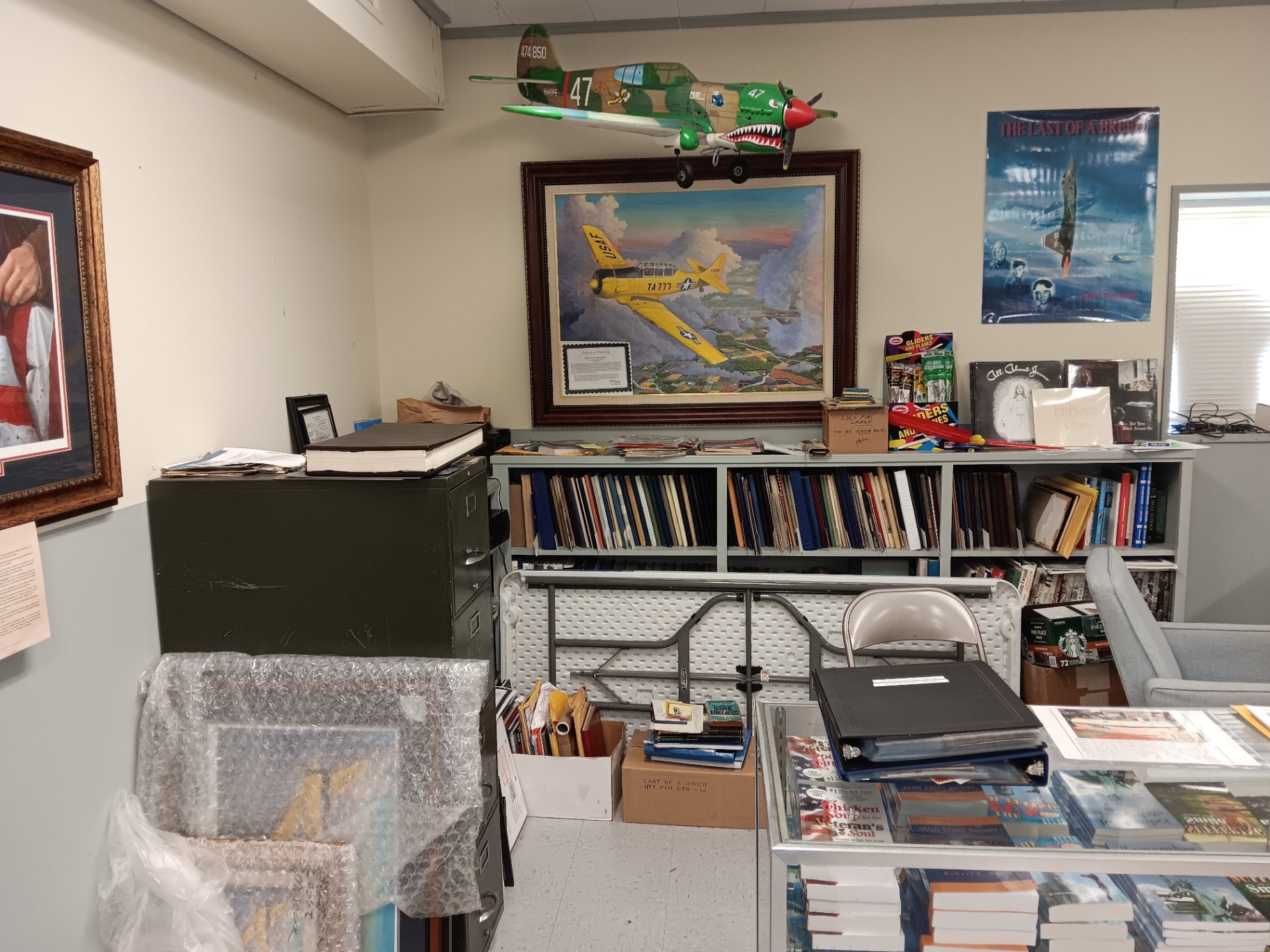 The reference library, a T-6 trainer painting, a shark-mouth P-40 overhead, and the poster for Errol's book “The Last of a Breed.”