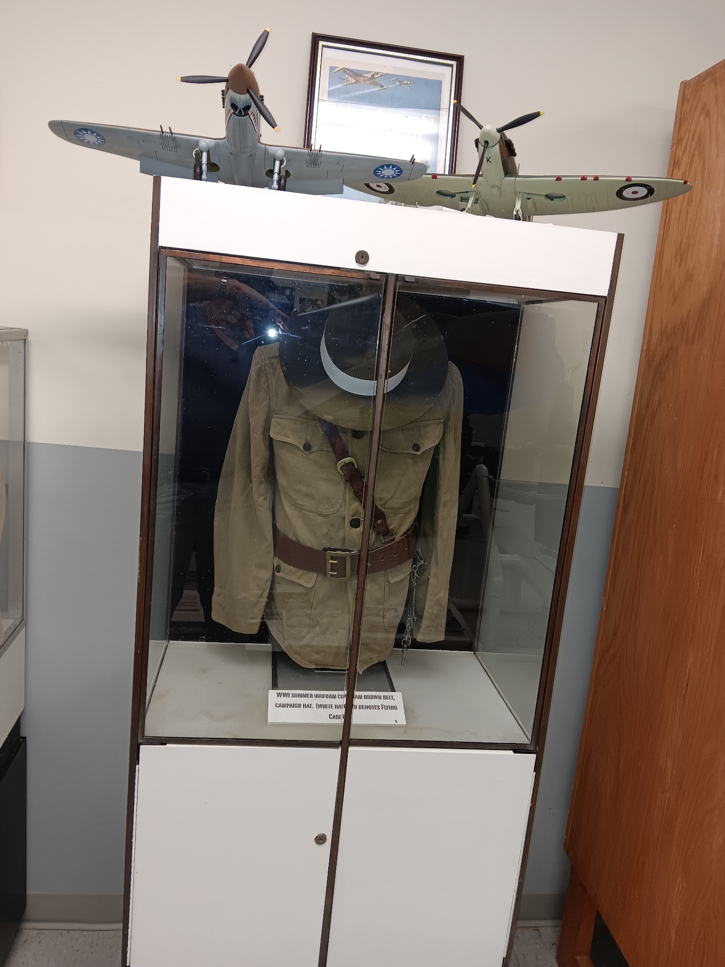 A WWI summer uniform coat with Sam Browne belt and campaign hat — the white hat band marks a Flying Cadet — under hand-built Spitfire and Flying Tigers models.